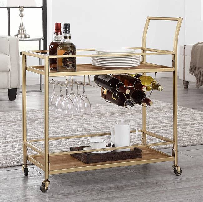 the gold bar cart holding glasses, bottles, plates, and a tea set