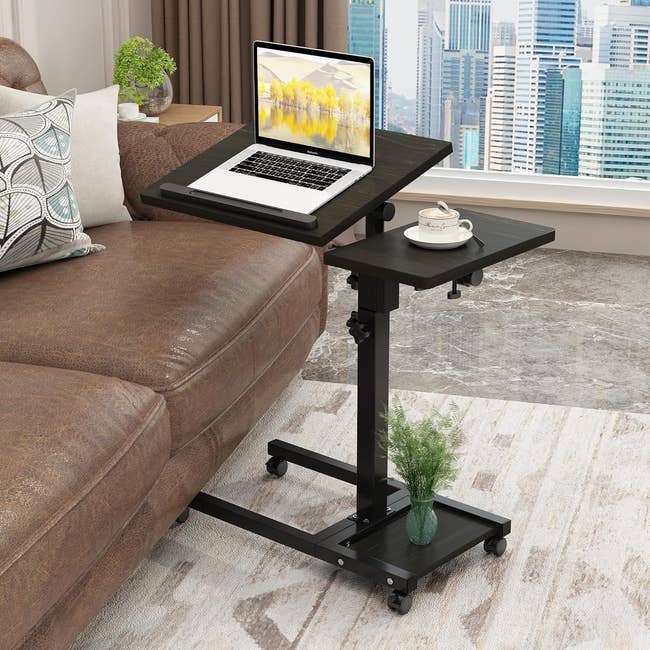 Adjustable laptop stand on wheels beside a sofa with a laptop, mug, and plant, designed for convenience and flexible use in living spaces
