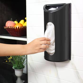 A person is using a black wall-mounted grocery bag dispenser in a kitchen setting