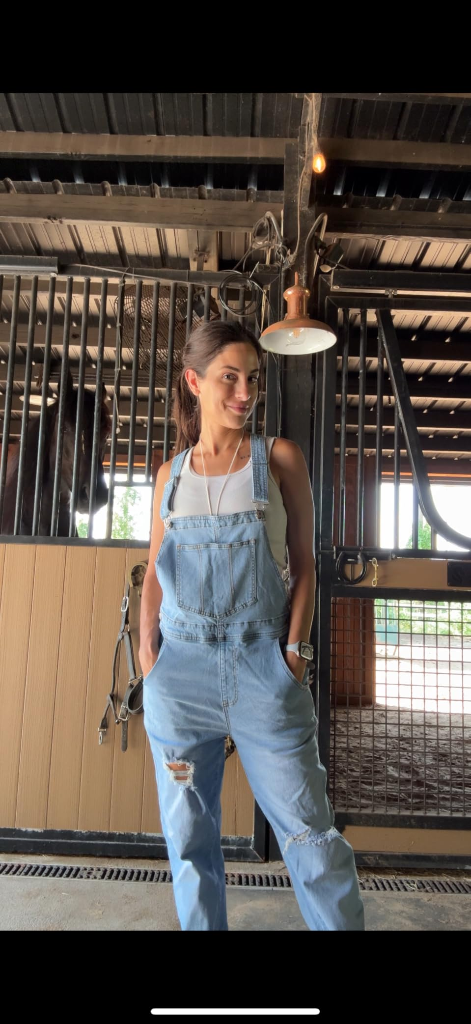 A idiosyncratic   stands successful  a unchangeable  wearing distressed denim overalls and a achromatic  top, with a equine  disposable   down  them successful  the stall