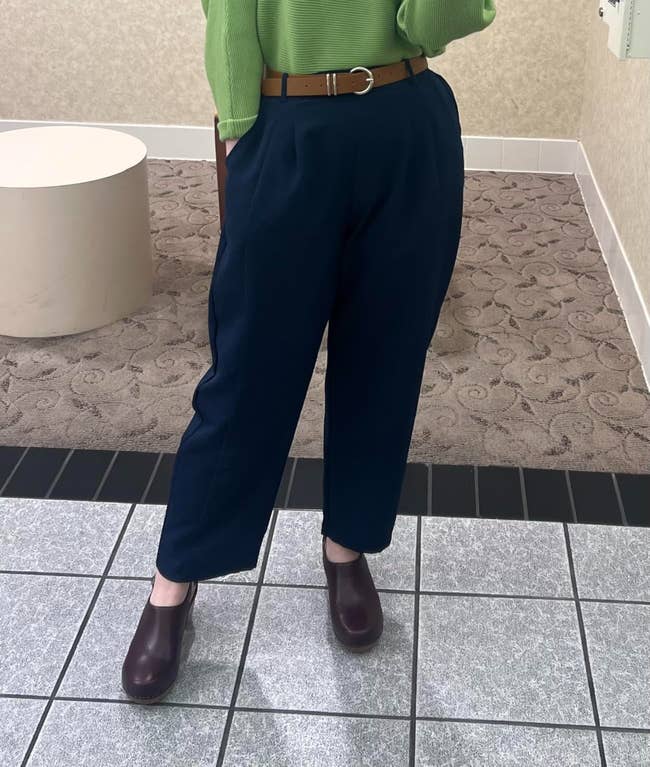 Person successful a greenish apical with bluish pleated pants, brownish belt, and burgundy shoes, lasting connected a tiled floor