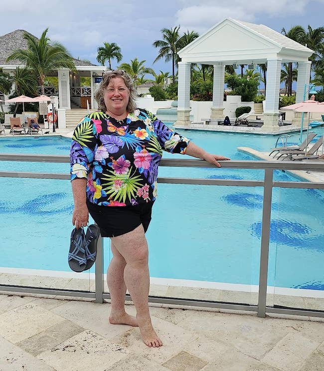 Person in tropical print shirt and shorts next to a pool, smiling, possibly on vacation
