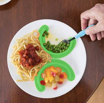 Plate with sectioned greenish  dividers holding pasta with herb  sauce, peas, and mixed fruit. Hand utilizing blue-handled spoon