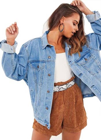 A person models a casual outfit with an oversized denim jacket, white tank top, and high-waisted brown corduroy shorts, accessorized with large hoop earrings
