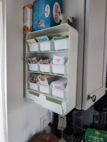 Wall-mounted organizer with various tea boxes and bags neatly arranged in compartments, beside kitchen cabinets