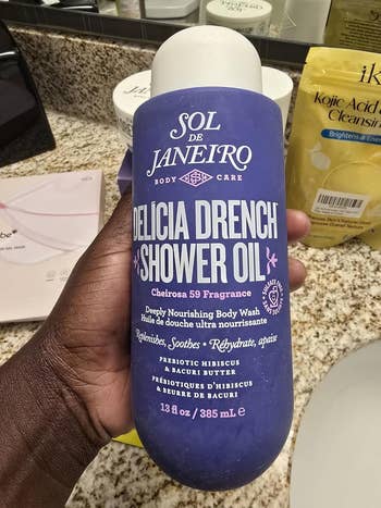 Person holding a bottle of Sol de Janeiro Delicia Drench Shower Oil on a bathroom counter