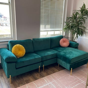 reviewer photo of the green velvet sofa in their house with two pincushion throw pillows in yellow and pink