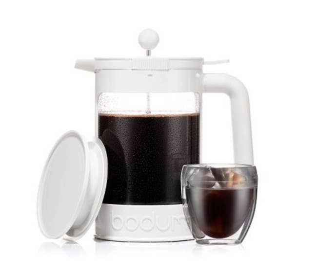 White cold brew maker with press lid and airtight circular lid next to glass of iced coffee on a white background