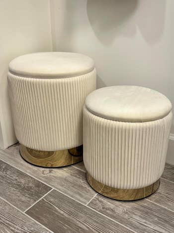 Two modern   cream-colored, pleated stools with golden  bases are displayed connected  a wood-tiled floor
