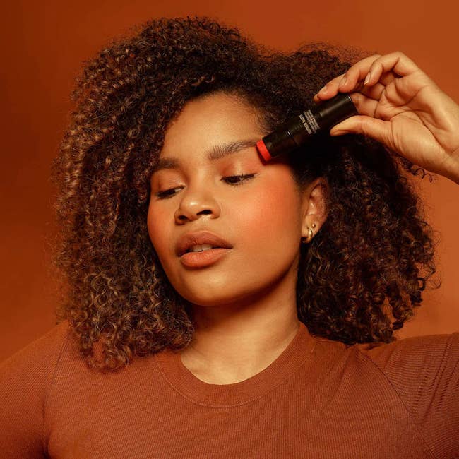 model holding the nudestix with it applied to their eyelids, cheeks, and lips, looking rosy and flushed