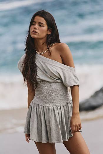 Model on a beach wearing an off-shoulder, short-sleeve, smocked gray dress with a gold necklace, looking relaxed with ocean waves in the background