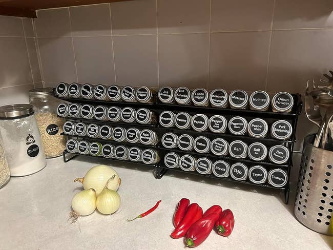 two spice racks with labeled jars on a kitchen counter, alongside onions and red peppers