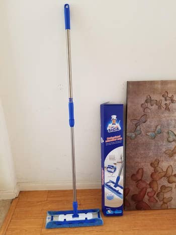 A STIGA flat floor mop with a microfiber pad next to its product box, leaning against an indoor wall