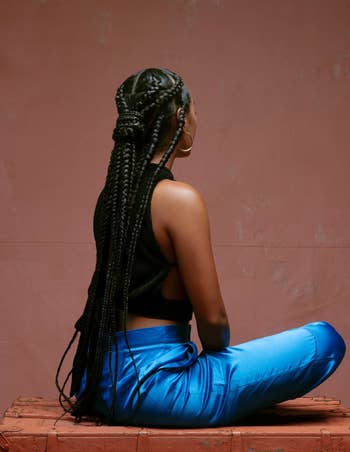 Person seated connected a box, facing away, with intricate braided hairstyle, wearing a sleeveless apical and shiny pants