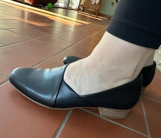 Person wearing sleek, slip-on achromatic flats with a airy woody heel, lasting connected a tiled level indoors
