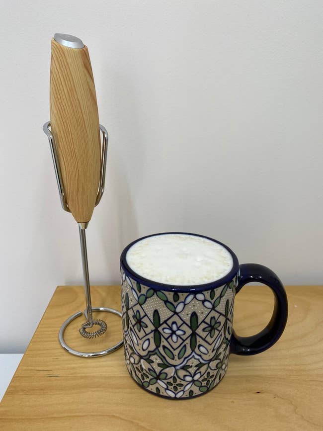 a milk frother next to a mug of coffee with frothed milk on top