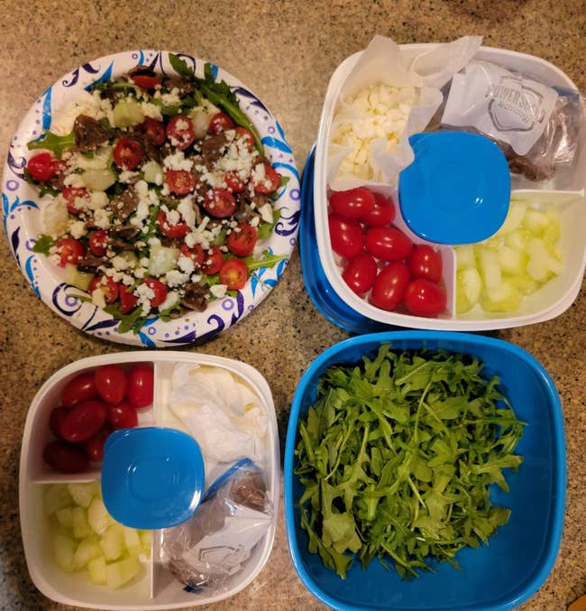 Four containers on a counter with salad ingredients: mixed greens, tomatoes, cucumbers, and feta cheese