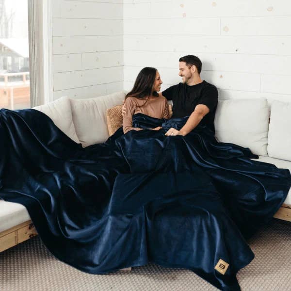 Couple sitting on a white sofa, wrapped in a large, luxurious blanket, smiling at each other