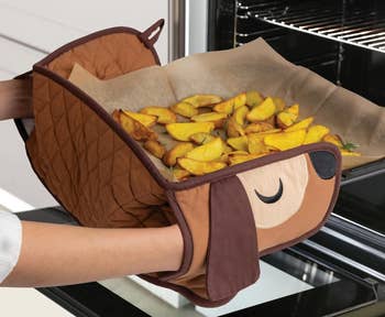 Oven mitts shaped like a dog's face are used to take out a tray of roasted potato wedges from an oven