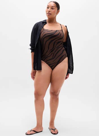 Person successful a zebra-print swimsuit and unfastened achromatic shirt, wearing flip-flops. They basal confidently against a plain background