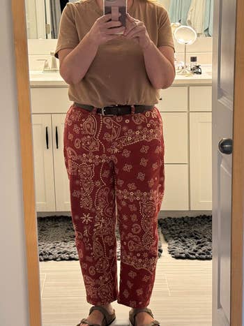 Person taking a reflector selfie wearing patterned pants, a plain shirt, and sandals successful a bath setting
