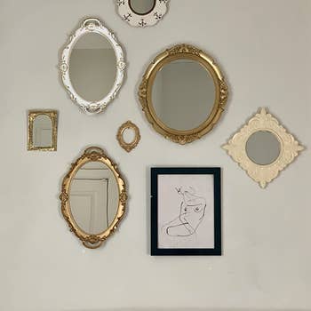 the white and gold versions of the mirror hung on a wall next to art and other mirrors as a sort of gallery wall
