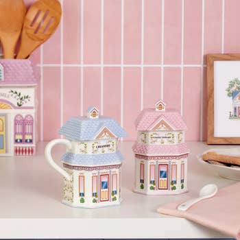 House-shaped ceramic jars with roof lids, styled like a creamery and confectionery, sit on a kitchen counter next to baking utensils