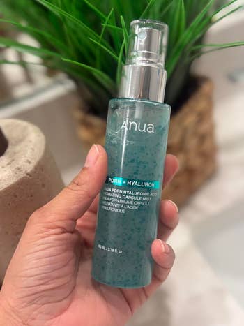 Hand holding a bottle of Anua PDRN Hyaluronic Acid Hydrating Capsule Mist, with a plant blurred in the background