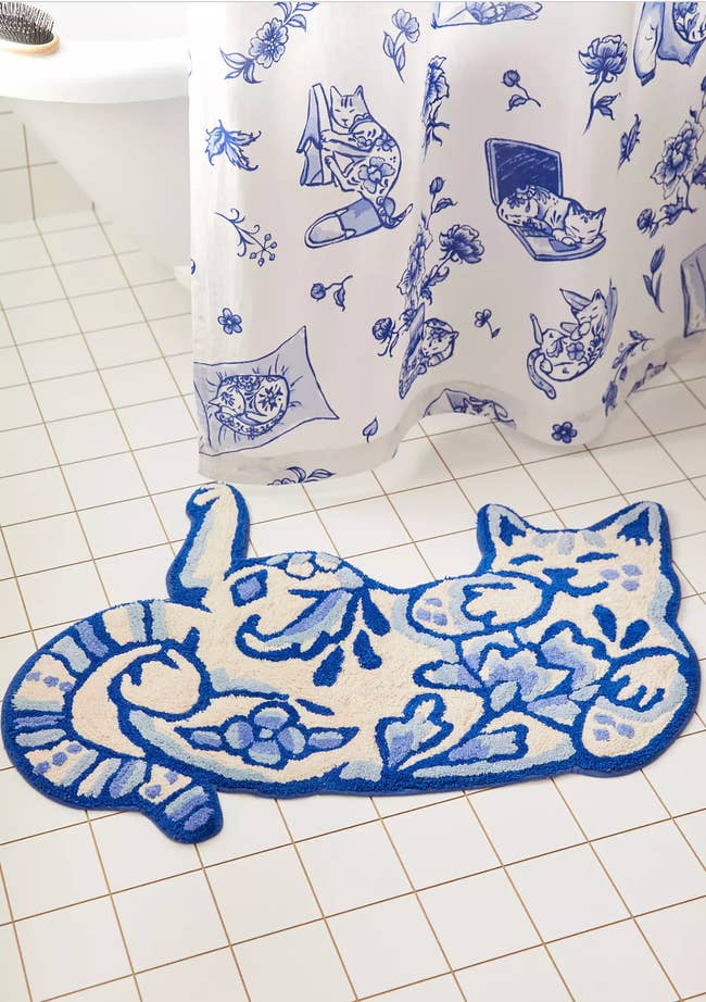 Cat-shaped bath mat with floral design on a white tiled floor, near a shower curtain with similar patterns. Ideal for bathroom decor