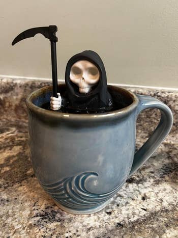 Grim Reaper figurine sitting in a coffee mug with a wave design, holding a scythe