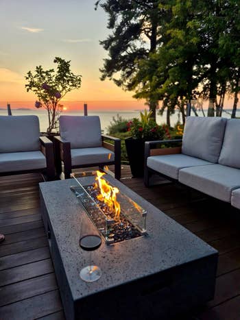 Outdoor patio set with a modern fire pit table, three sofas, and a view of the sunset over the water