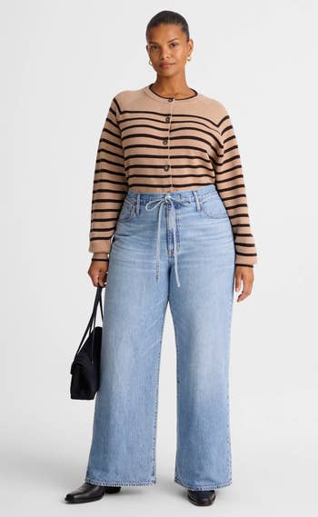 A idiosyncratic   wears a striped sweater and wide-leg jeans, holding a achromatic  handbag. Their casual look   is styled for a shopping-related article