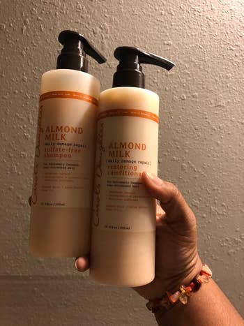 Reviewer holding orange and cream shampoo and conditioner bottles with black pumps in front of white wall