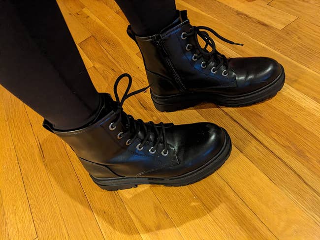 Person wearing stylish achromatic combat boots connected a woody floor, paired with achromatic pants