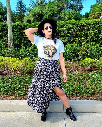 reviewer in a graphic tee, floral skirt, boots, and a wide-brimmed hat, posing with one hand on hip