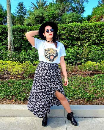 reviewer in a graphic tee, floral skirt, boots, and a wide-brimmed hat, posing with one hand on hip