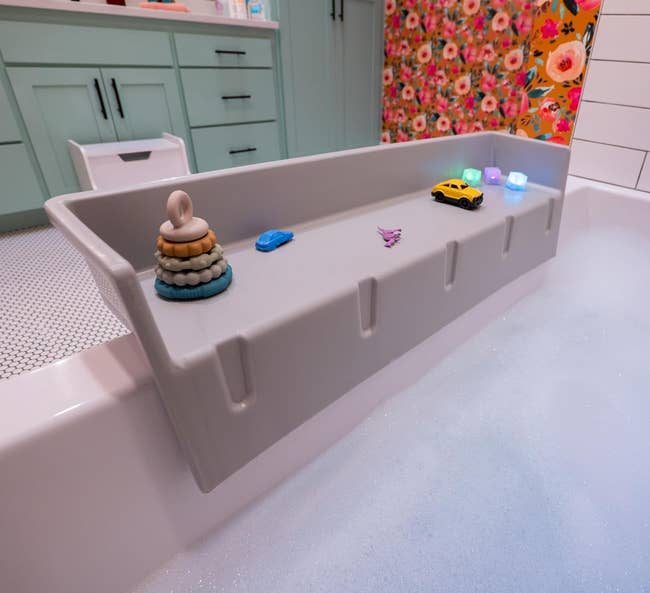 The gray bathtub topper on the lip of the tub with toys on it for play