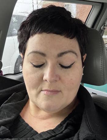 Person with short hair and winged eyeliner sits in a car with eyes closed, wearing a textured black top