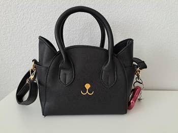 Black handbag with gold embroidery, top handles, and a shoulder strap, featuring a small pink charm. Ideal for stylish everyday use