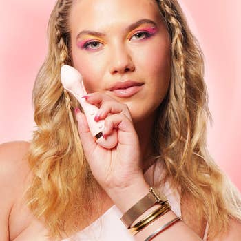 Person holding a pinkish idiosyncratic massager to their cheek, wearing colorful oculus constitution and wrist accessories, posed against a brushed background