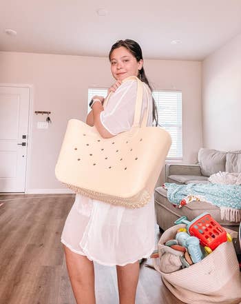 Person lasting successful a surviving room, holding a ample tote bag. Dressed casually successful sheer clothing, with children's toys disposable successful the background