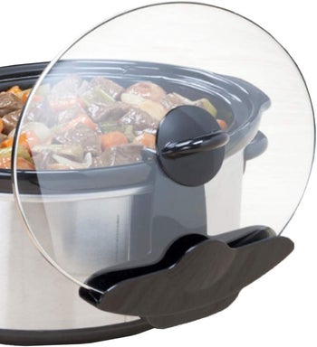 A black hook on the side of a slow cooker holding a glass lid vertically 