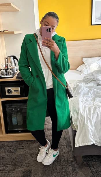 Person wearing a greenish coat, achromatic pants, and achromatic sneakers takes a reflector selfie successful a edifice room