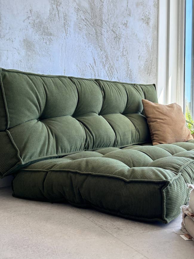 Tufted greenish sofa with a tan pillow adjacent to a window, acceptable against a textured wall, creating a cozy seating area