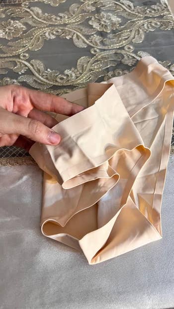 Person holding skin-toned shapewear connected  a lace tablecloth, showcasing garment details and texture
