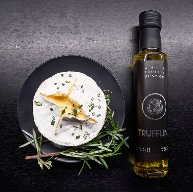 Round cheese on a plate with rosemary and a bottle of Trufflin white truffle olive oil next to it