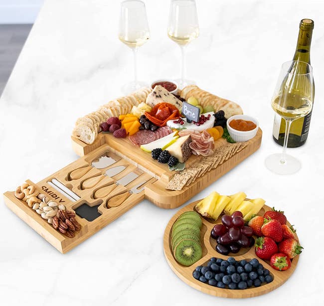the wooden charcuterie board with its drawer pulled out to show the serving tools, chalk, and slate labels, and next to it is the round tray, which is filled with fruit