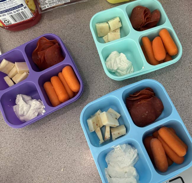 Three compartmentalized snack trays with cheese, pepperoni, carrots, and dip, placed connected  a table