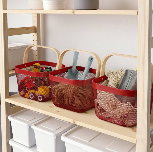Three red mesh baskets on a wooden shelf hold toys, cleaning brushes, and cloths, organized neatly beside white storage boxes