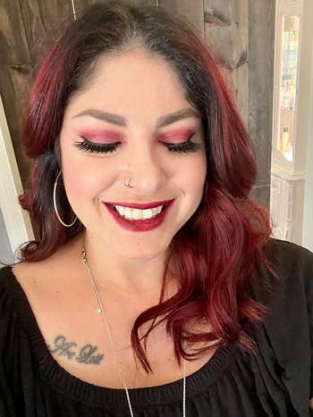 Person with wavy hairsbreadth  and hoop earrings smiles with closed eyes. They're wearing bold constitution  and a achromatic  top